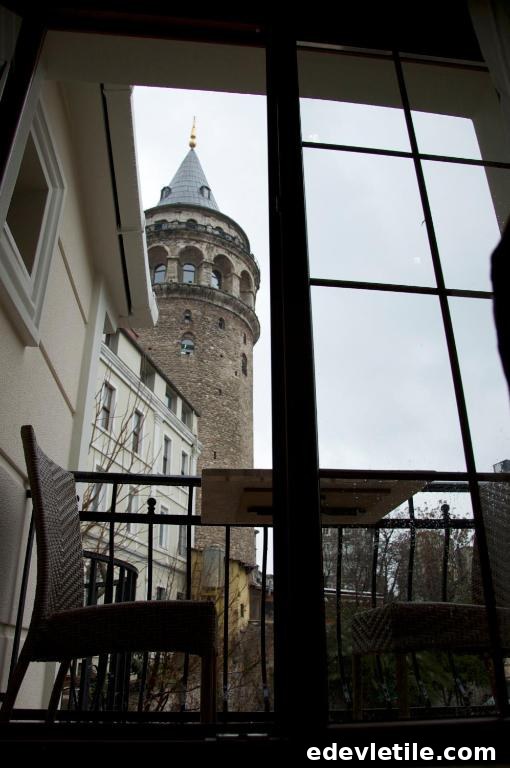 View of the Galata Tower on a cloudy day, framed by a balcony with chairs outside a hotel room window.