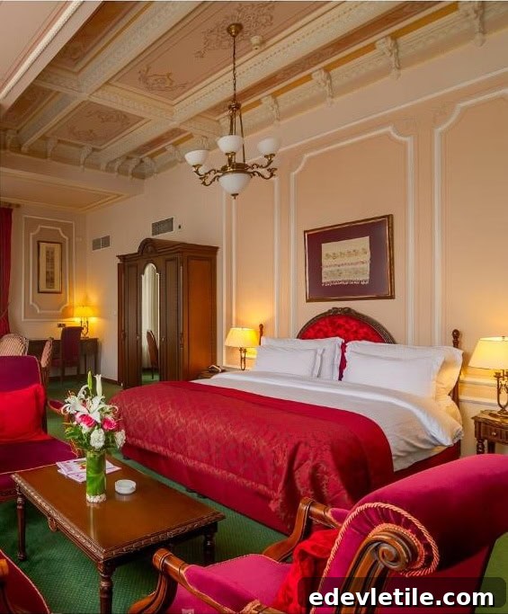Grand, vintage-style hotel room featuring a red velvet bedspread, carved wood furniture, and ornate ceiling molding.