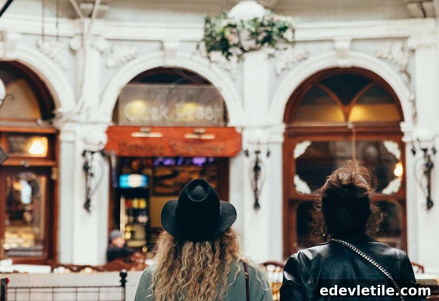 Two women viewed from behind walking through a grand indoor passage (Cicek Pasaj) in Istanbul, lined with arched windows and ornate details reminiscent of Beyoğlu’s Belle Epoque architecture.
