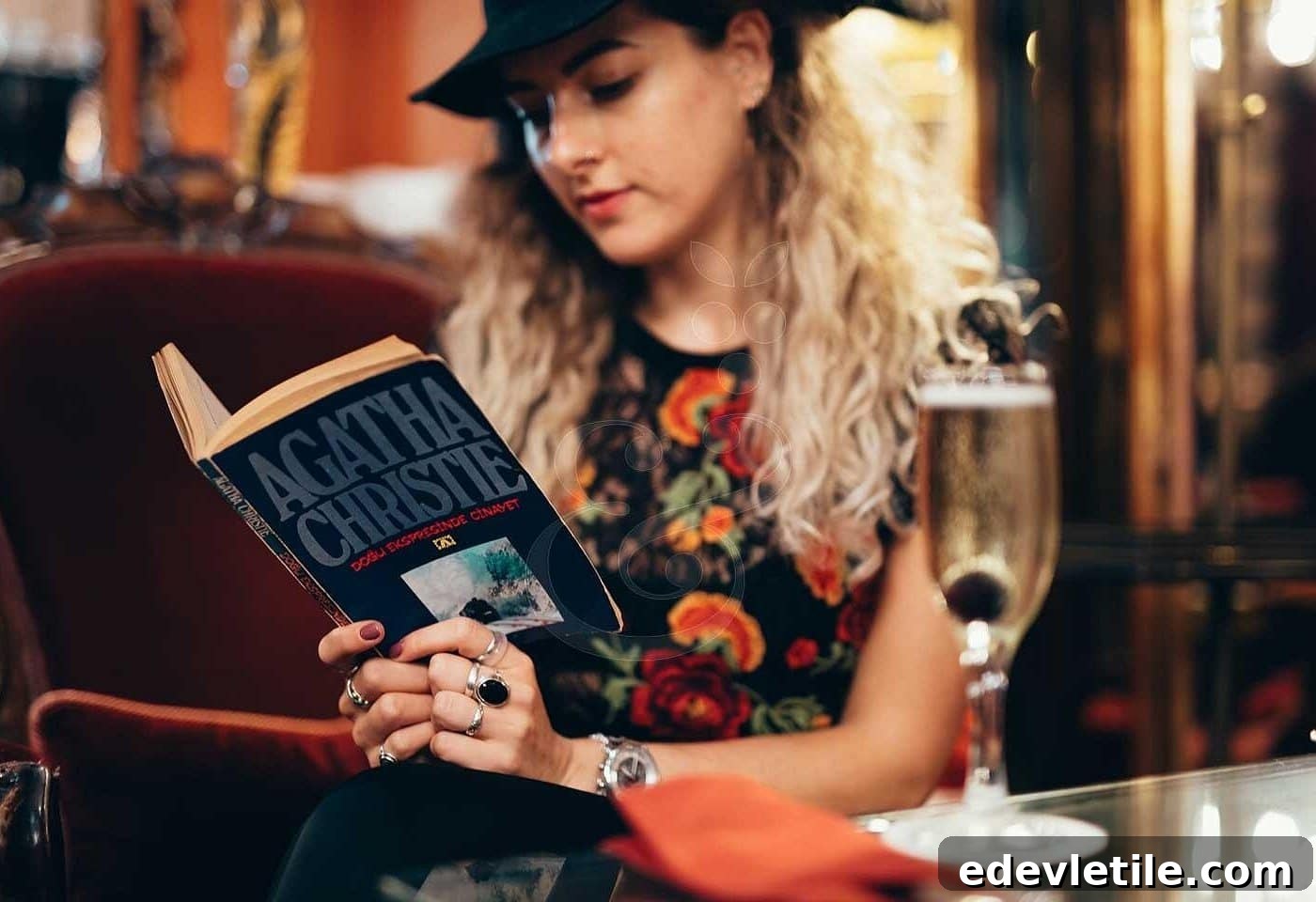 Woman dressed in a floral top and wide-brim hat reads an Agatha Christie novel while seated in a plush, vintage-style lounge with a glass of sparkling wine on the table beside her.