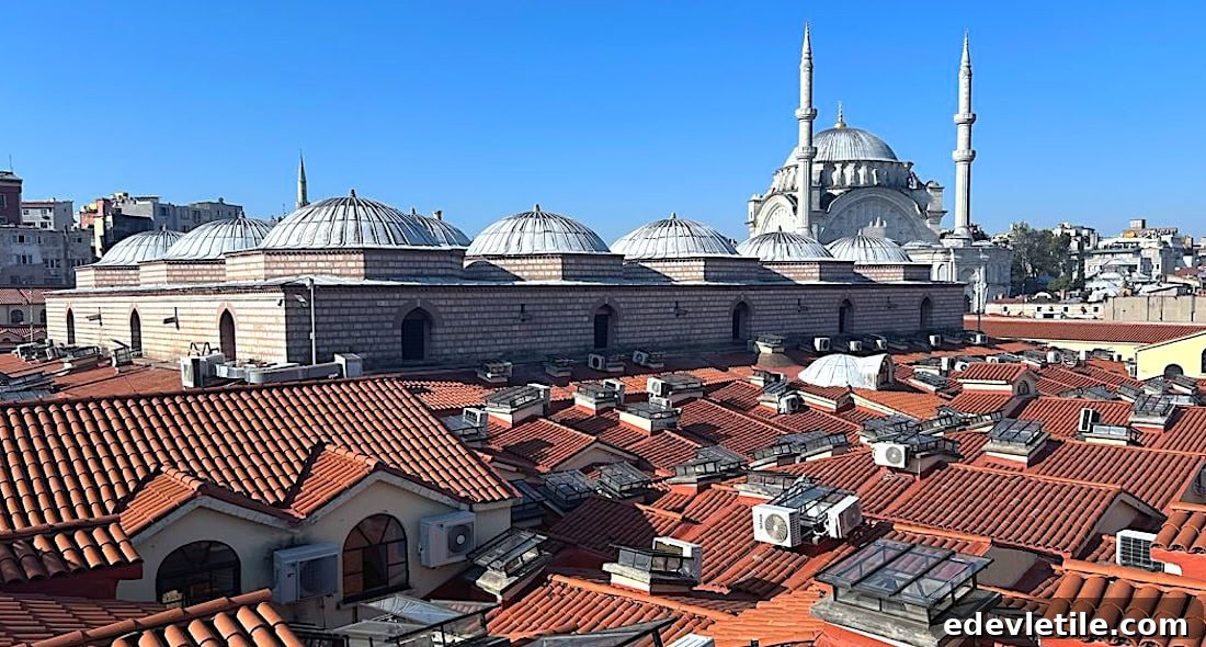 Rooftop view of Istanbul’s Grand Bazaar, with rows of red terracotta tiles and scattered skylights, backed by the domed and minareted mosque under a clear blue sky.
