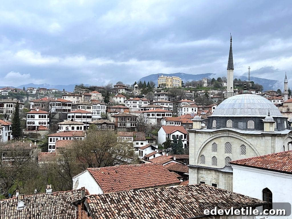 Panoramic view of Safranbolu’s red-roofed Ottoman houses and historic mosque dome, under a cloudy sky with rolling hills in the background.