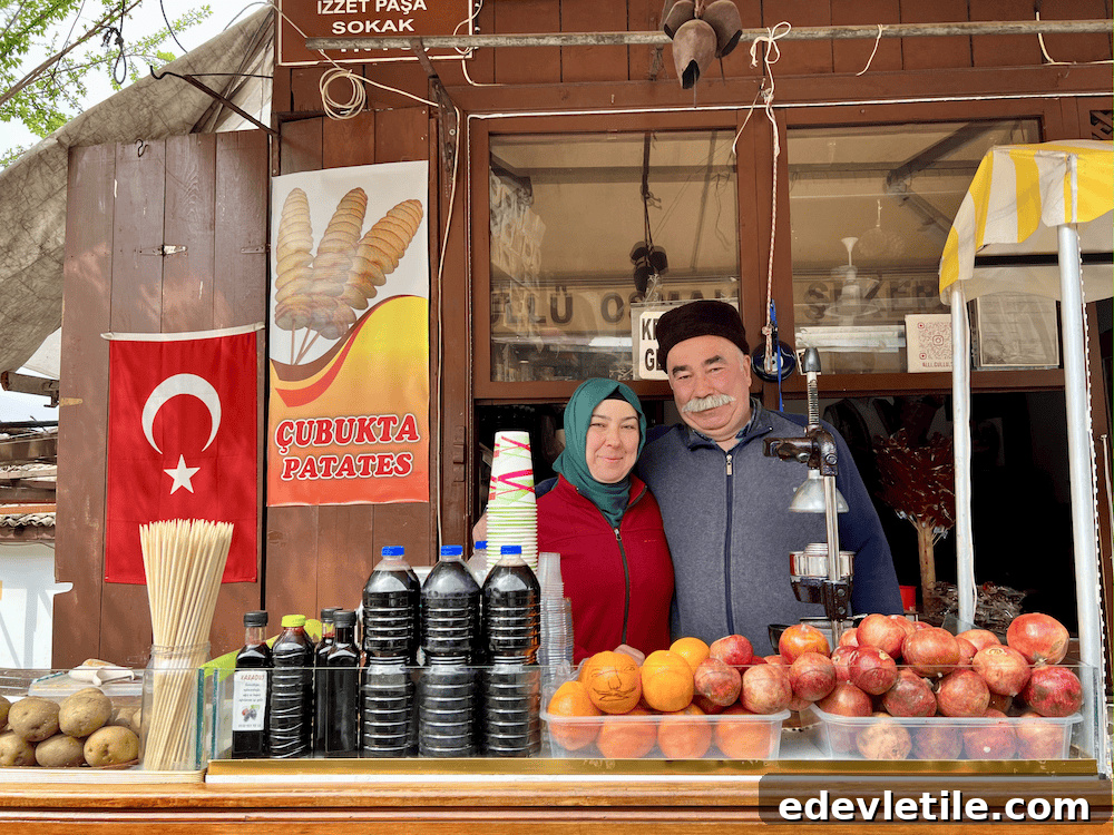 Smiling couple at a traditional juice stand in Safranbolu, surrounded by pomegranates, oranges, and syrup bottles, with a Turkish flag and sign for spiral potatoes.
