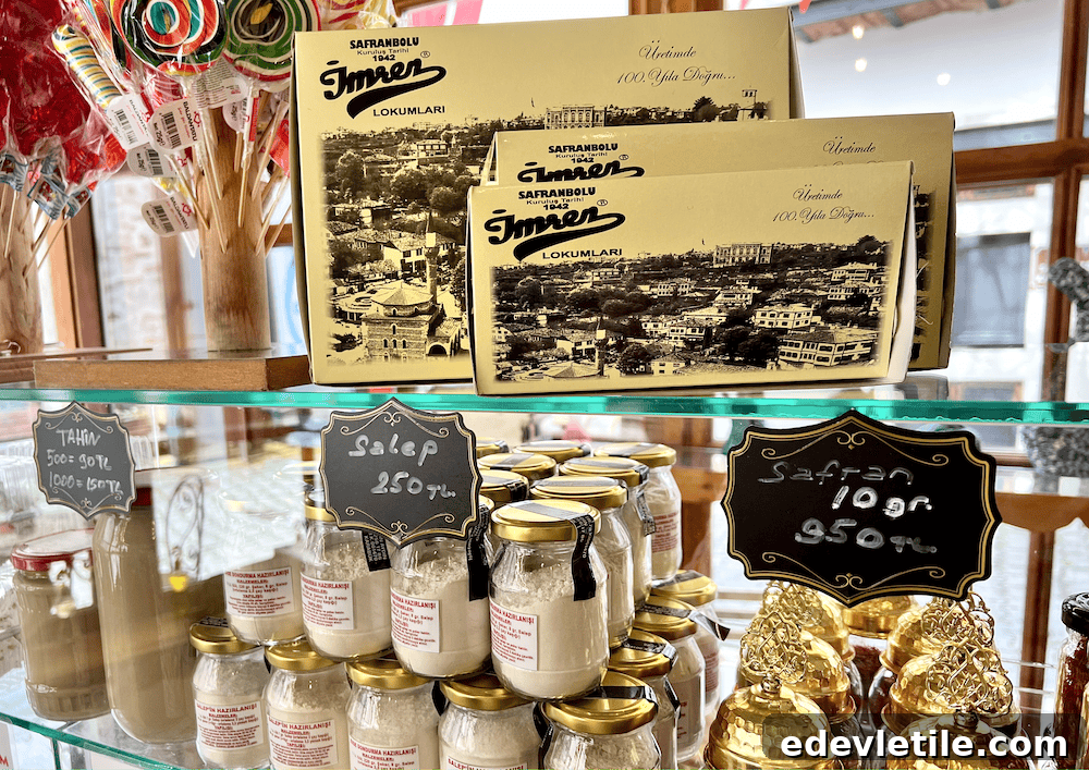 Glass shelf featuring jars of salep and tahini, boxes of lokum, golden saffron tins, and colorful lollipops in a traditional Safranbolu store.