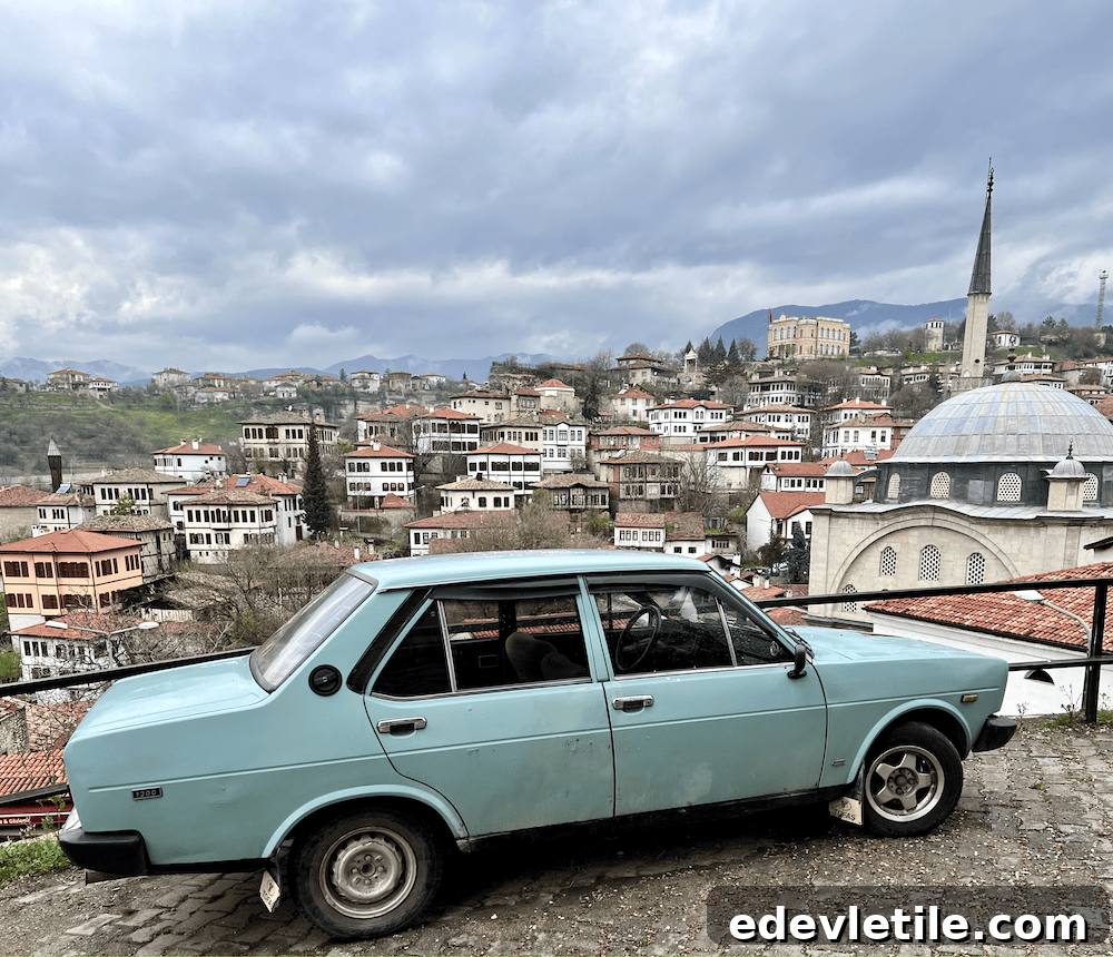 Light blue classic car parked on a cobbled hill overlooking Safranbolu’s red-roofed homes, mosque, and lush green hillside under a cloudy sky.