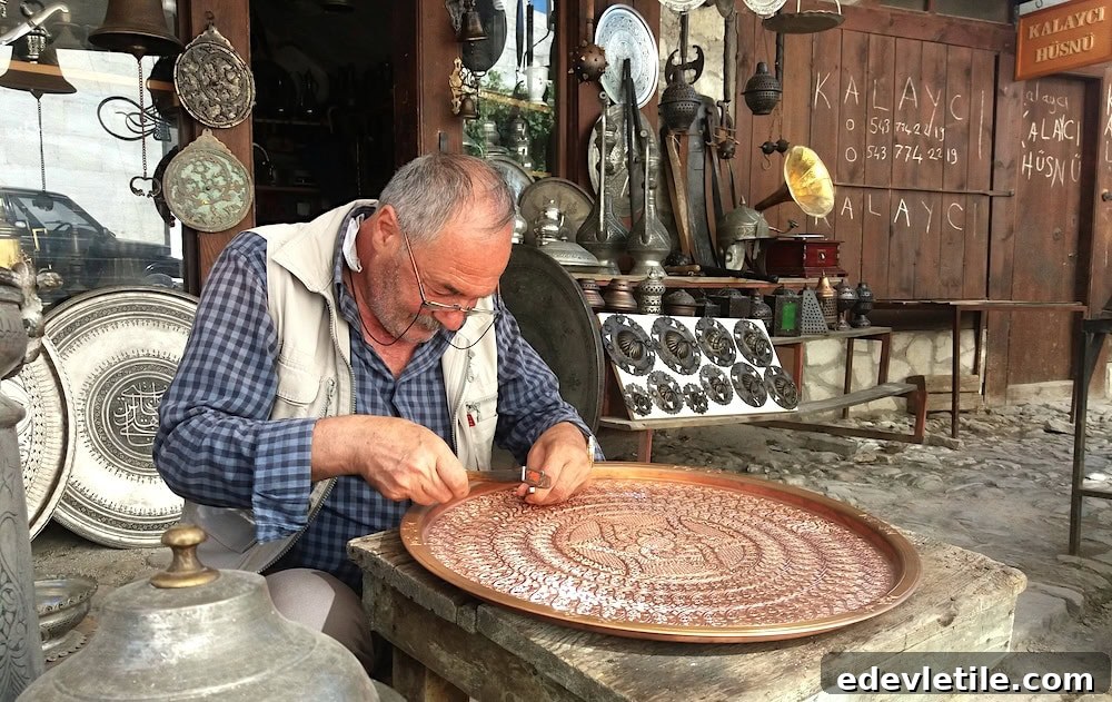 Elderly artisan seated outside his shop in the Safranbolu bazaar, meticulously hand-engraving an ornate copper tray surrounded by antique metal goods and hanging decorations.