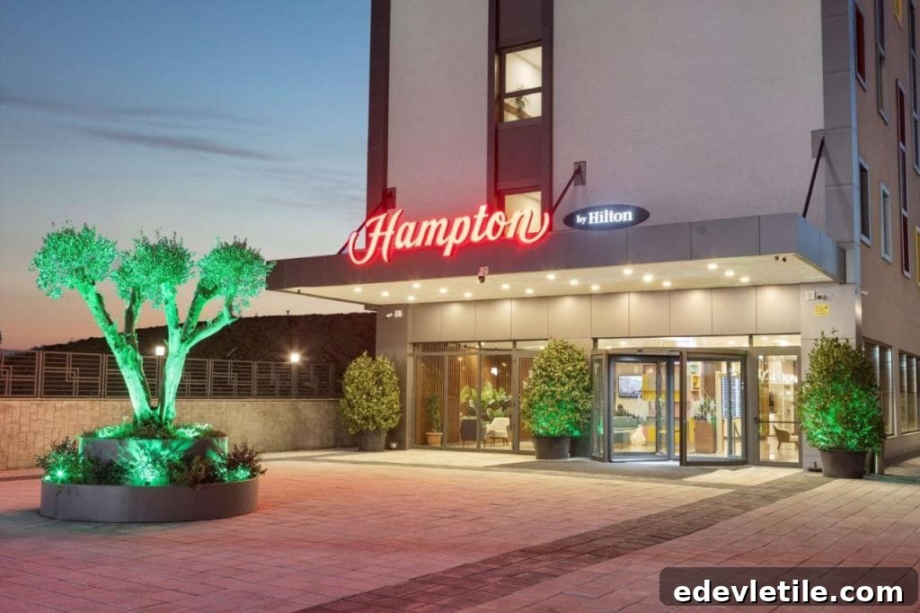 Exterior view of Hampton by Hilton Istanbul Arnavutköy at dusk, with glowing red signage and a tree lit in green.