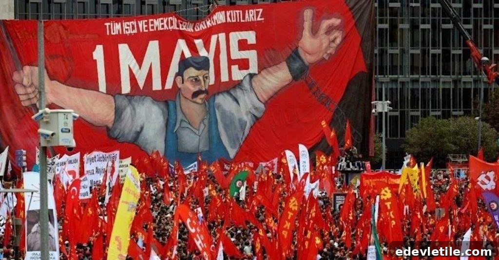 A large crowd holds red flags below a giant banner reading “1 Mayıs” with a worker illustration, celebrating International Workers’ Day in Turkey.