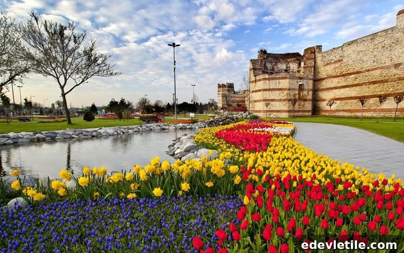 Tulip-lined pond near the ancient city walls of Istanbul, with neatly arranged red, yellow, and blue flowers curving along a stone path.