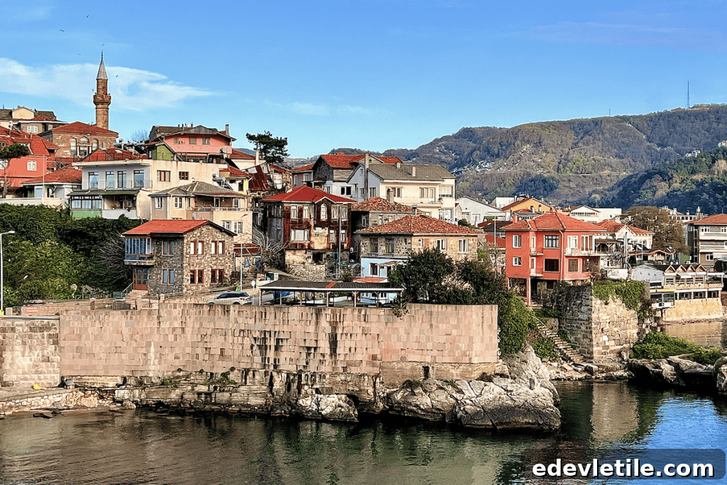 Historic homes with red rooftops and stone walls line the rocky coastline of Amasra, backed by lush spring hills under a clear sky.