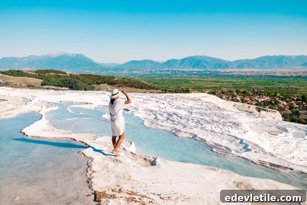 A woman in a white outfit and sunhat standing barefoot on the edge of a Pamukkale thermal pool, gazing at the valley below and distant mountains.