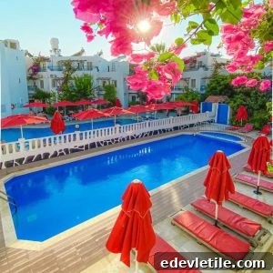 Bright hotel pool area surrounded by red sun loungers and umbrellas, with vibrant pink bougainvillea flowers in the foreground and white-washed buildings in the background.
