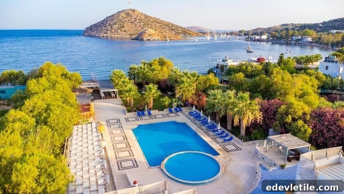 Aerial view of a serene hotel in Gümüşlük with a large pool surrounded by palm trees and blue loungers, overlooking a calm bay with boats and a small island.