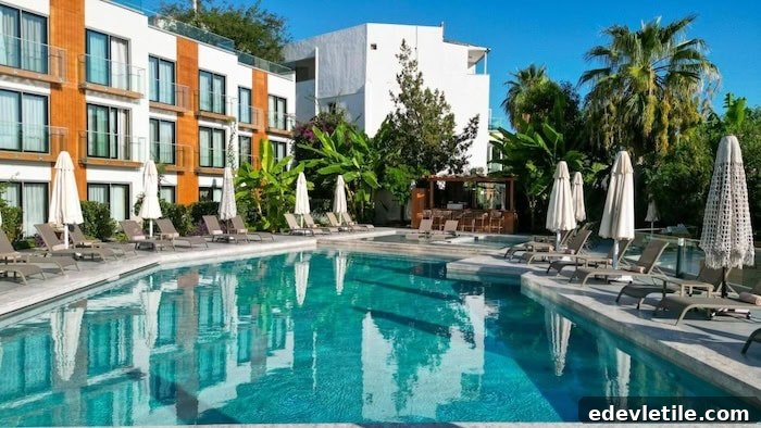 Modern boutique hotel in Gümüşlük with a turquoise pool, lined with sun loungers and surrounded by lush greenery and a mix of white and wood-accented buildings.