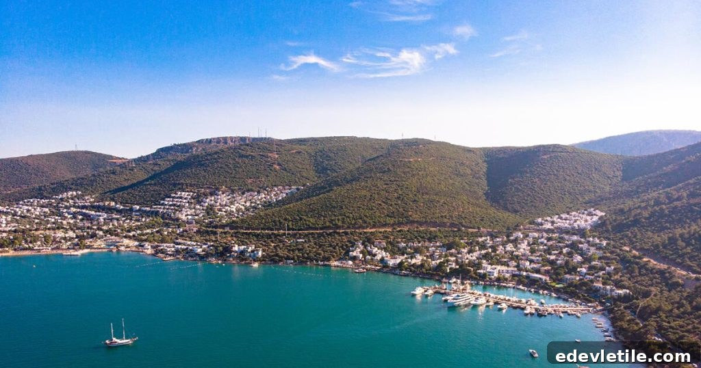 Aerial shot of Gündoğan Bay showing whitewashed homes nestled into forested hillsides, a marina, and turquoise waters under a bright blue sky.