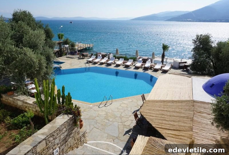 Aerial view of a coastal hotel in Torba with a blue geometric pool, sun loungers, and panoramic views of the sea and surrounding hills.
