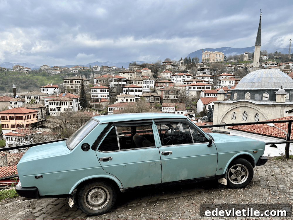 A light blue vintage car parked on a cobbled street overlooking the hillside town of Safranbolu, with traditional Ottoman houses and a mosque in the background.