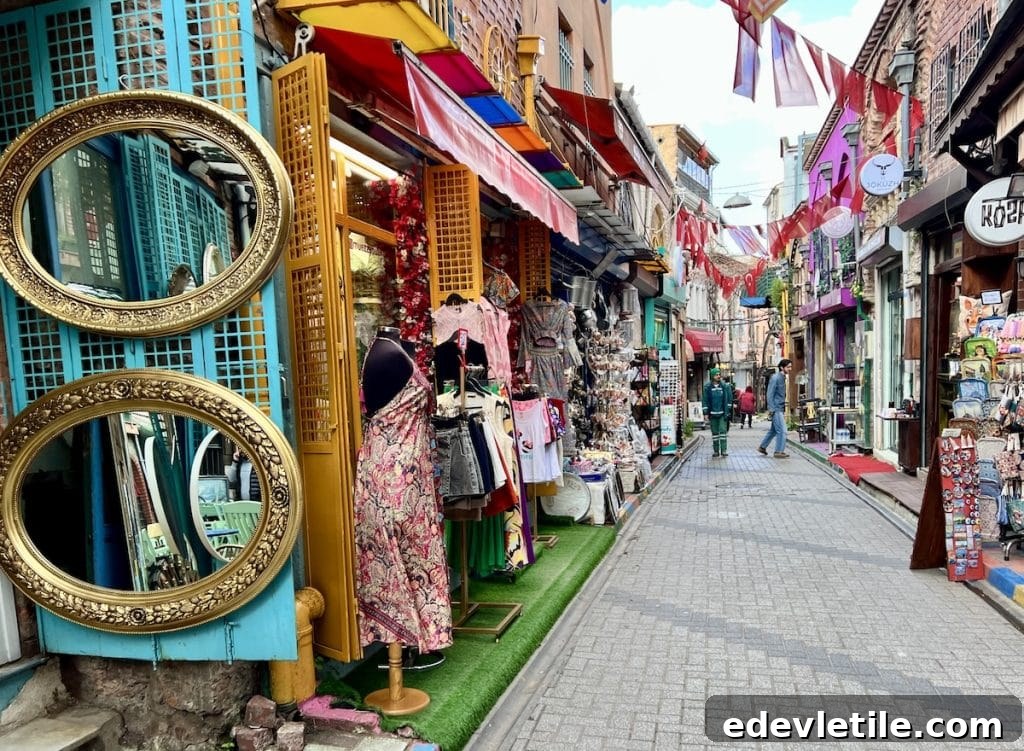 Narrow pedestrian alley in Balat lined with colorful shops selling dresses, souvenirs, and home goods, with red and white flags and lanterns strung above.