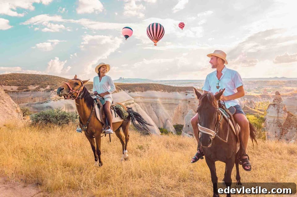 A couple riding horses through golden fields in Cappadocia, with scenic valleys and floating hot air balloons in the background.