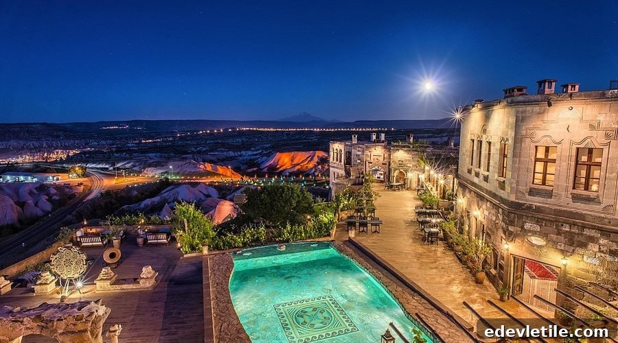 Scenic view of a boutique cave hotel with a lit-up pool and panoramic views of Cappadocia's dramatic landscape under a full moon.