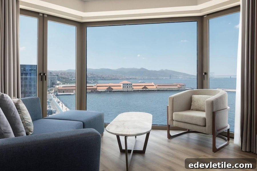 Sea view from a modern hotel room in Izmir.
Corner hotel suite with panoramic floor-to-ceiling windows overlooking the sea, a port building with a red roof, and distant hills.