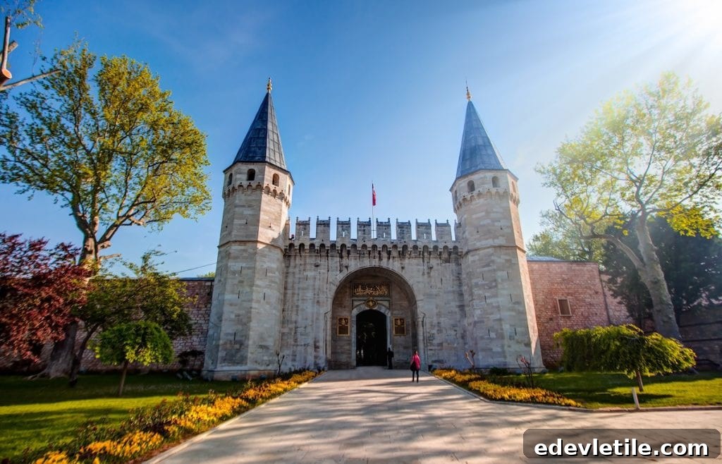 Grand main entrance to Topkapi Palace with pointed towers and stone walls, set against a bright blue sky and garden-lined path.