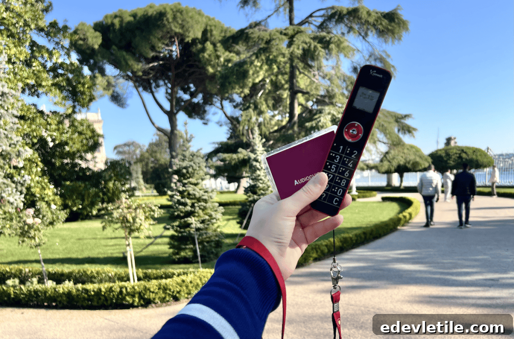 Visitor holding a ticket and audio guide for Dolmabahçe Palace in a garden area with trimmed trees and a view of the Bosphorus in the background.