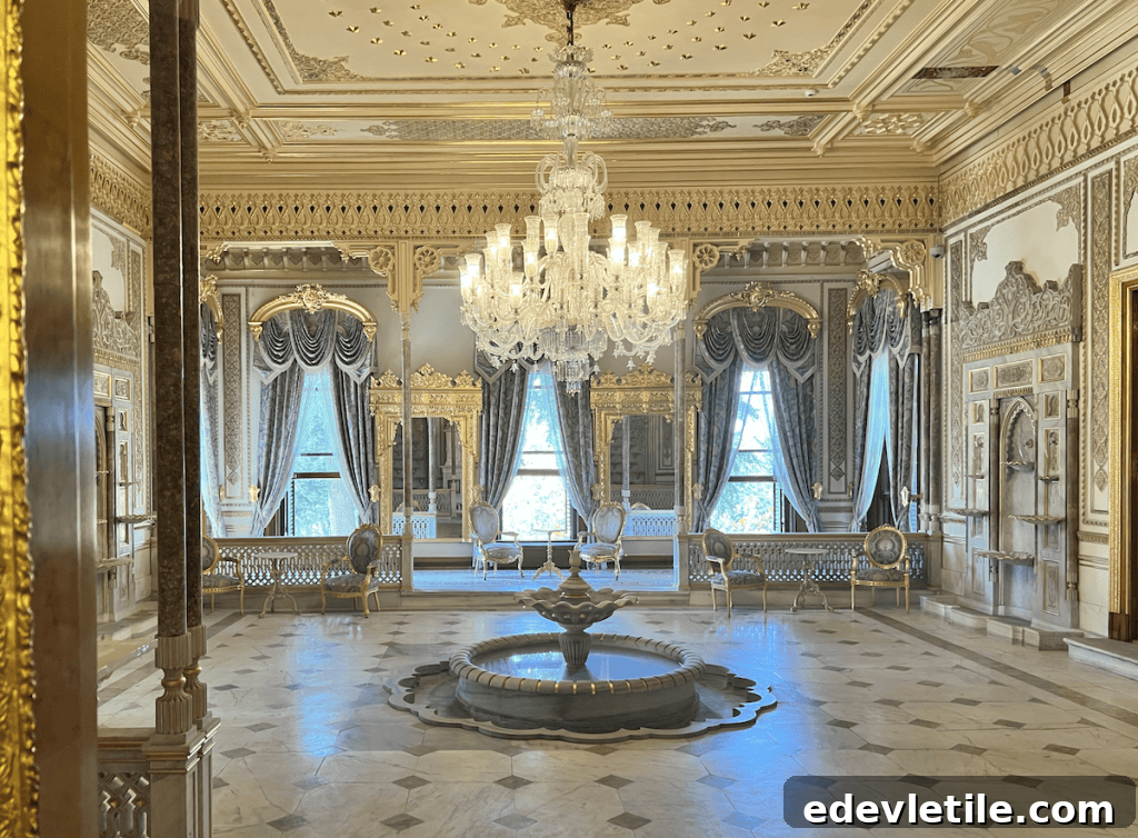 Luxurious palace room featuring a central marble fountain, crystal chandelier, ornate golden mirrors, and richly draped windows.