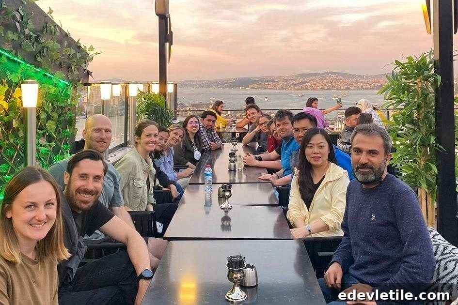 A large group of people sit at a long rooftop table at sunset, with views of the Bosphorus in the distance, enjoying a night food tour in Istanbul