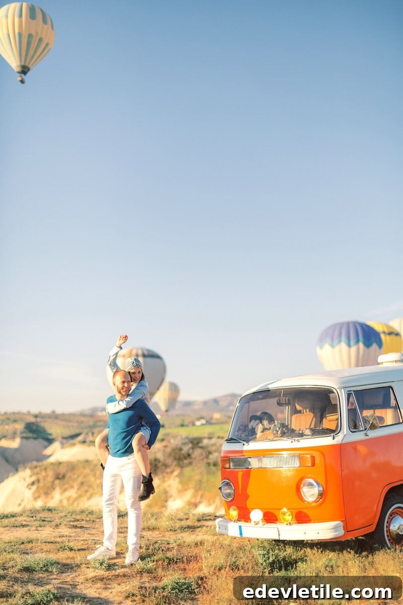 Man carrying a woman on his back beside a vintage orange van in Cappadocia, with hot air balloons rising in the distance on a clear morning.