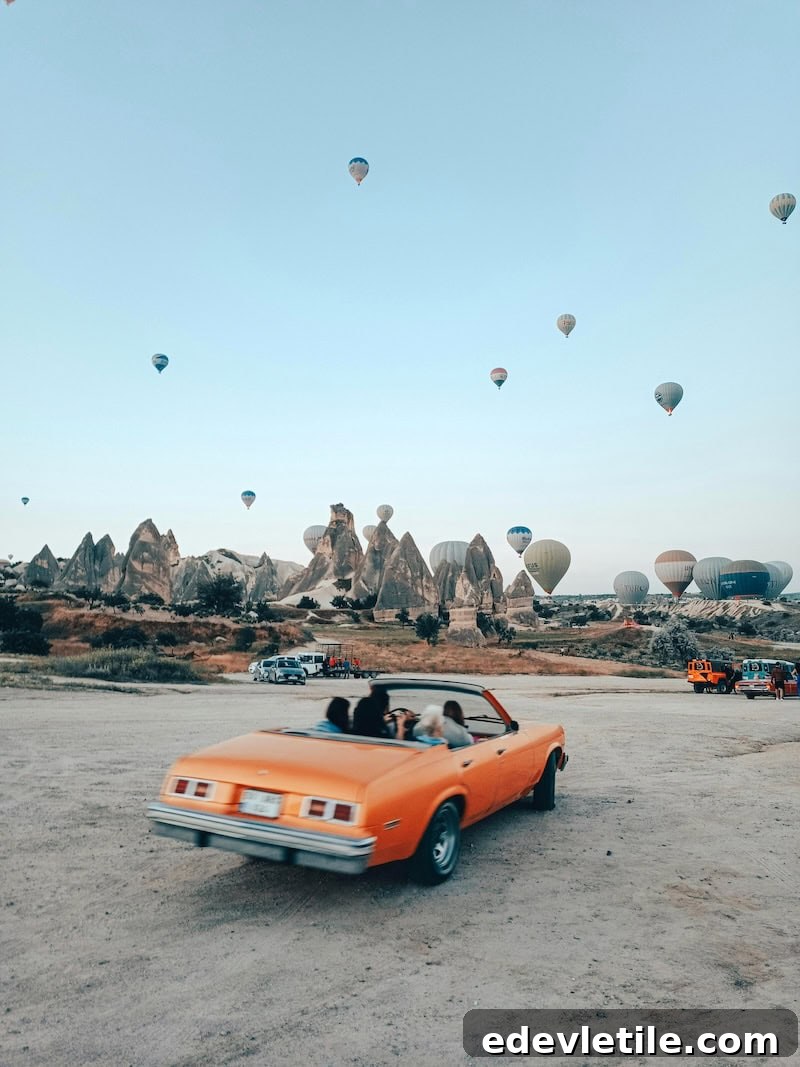 Bright orange convertible car driving through a sandy viewpoint in Cappadocia, with people inside and dozens of hot air balloons in the sky over jagged fairy chimneys.
