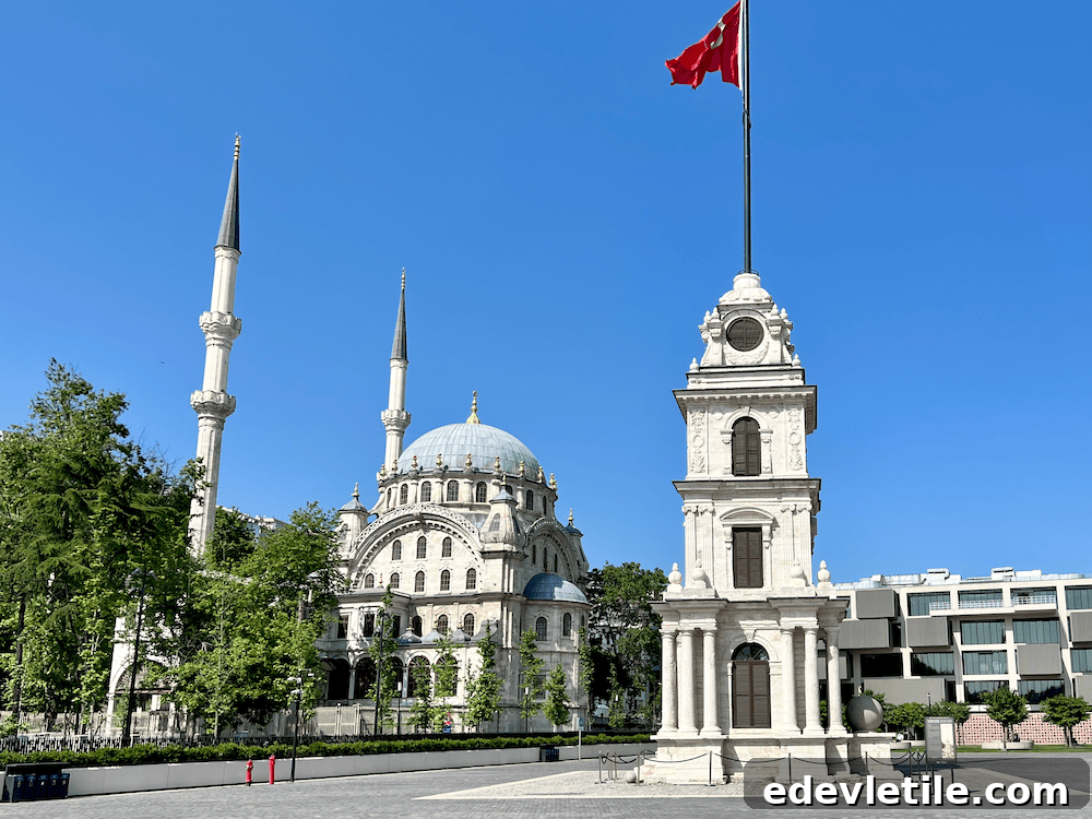 View of the ornate Nisretiye Mosque (Nisretiye Cami) and historic Tophane clock tower near Galataport Istanbul, with a Turkish flag flying high against a clear blue sky.