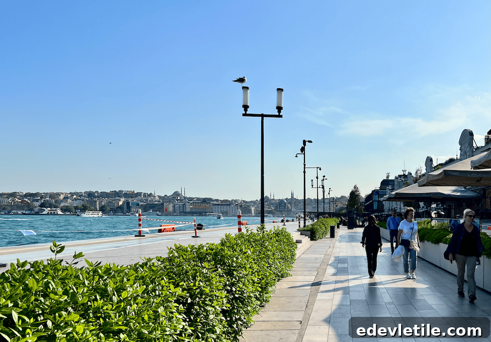 Seaside promenade at Galataport Istanbul with people walking along the Bosphorus waterfront, modern cafes to the right and historic cityscape across the water.