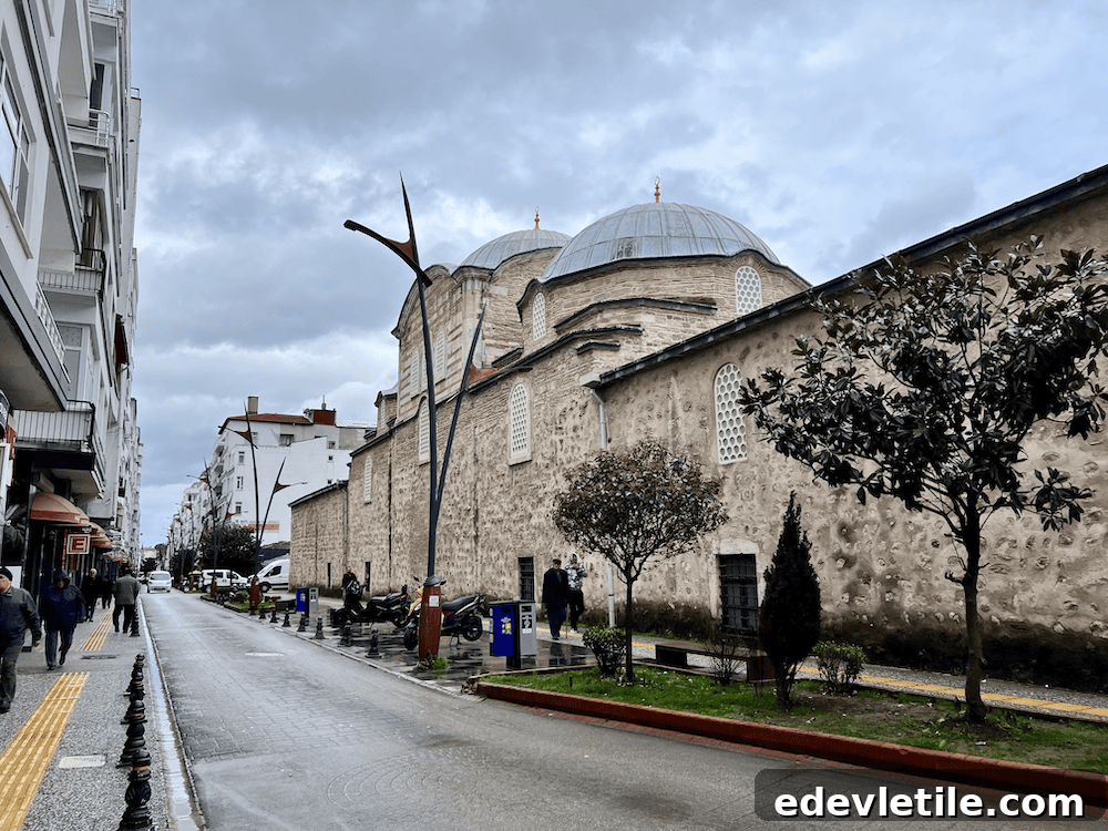 Street view of Sinop’s historic Alaaddin Mosque with its stone walls and domes, set along a quiet wet road lined with local buildings and trees.