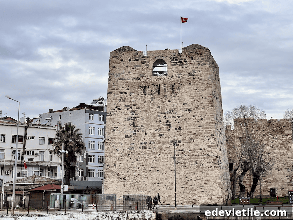 Large stone watchtower of the Sinop Fortress, flying a Turkish flag, with nearby residential buildings and a cloudy sky in the background.