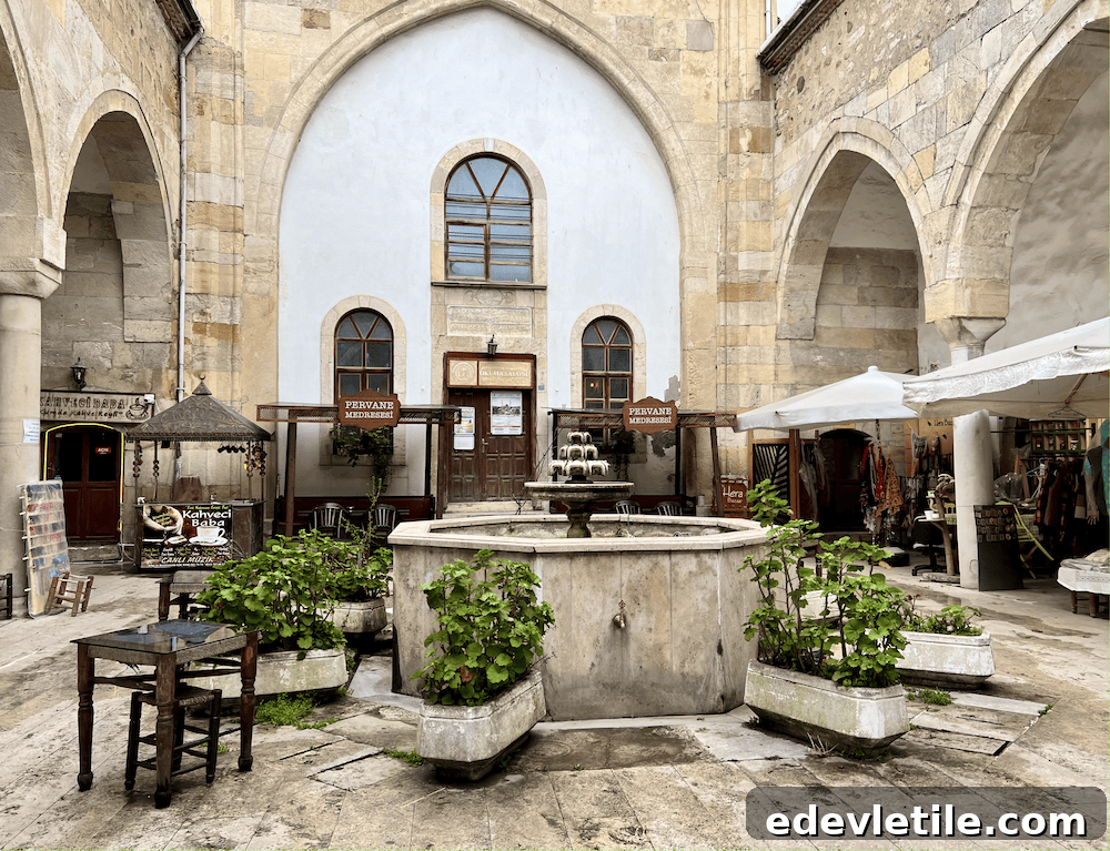 Courtyard of the historic Pervane Medrese in Sinop, Turkey, featuring a central stone fountain, arched stone walls, small shops, and seating areas surrounded by greenery.